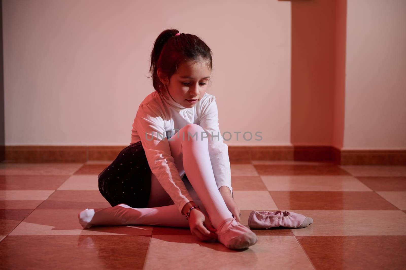 A young ballerina in a studio ties her ballet shoes, surrounded by warm, focused lighting, symbolizing dedication and the beauty of dance.