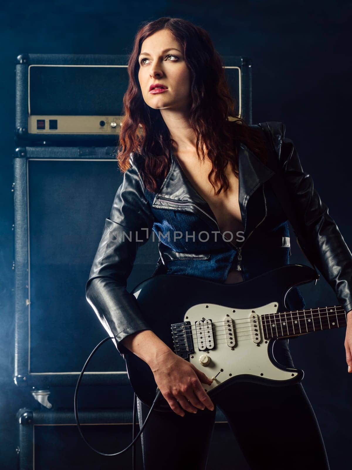 Beautiful young woman playing electruc guitar on stage in front of large amplifier.