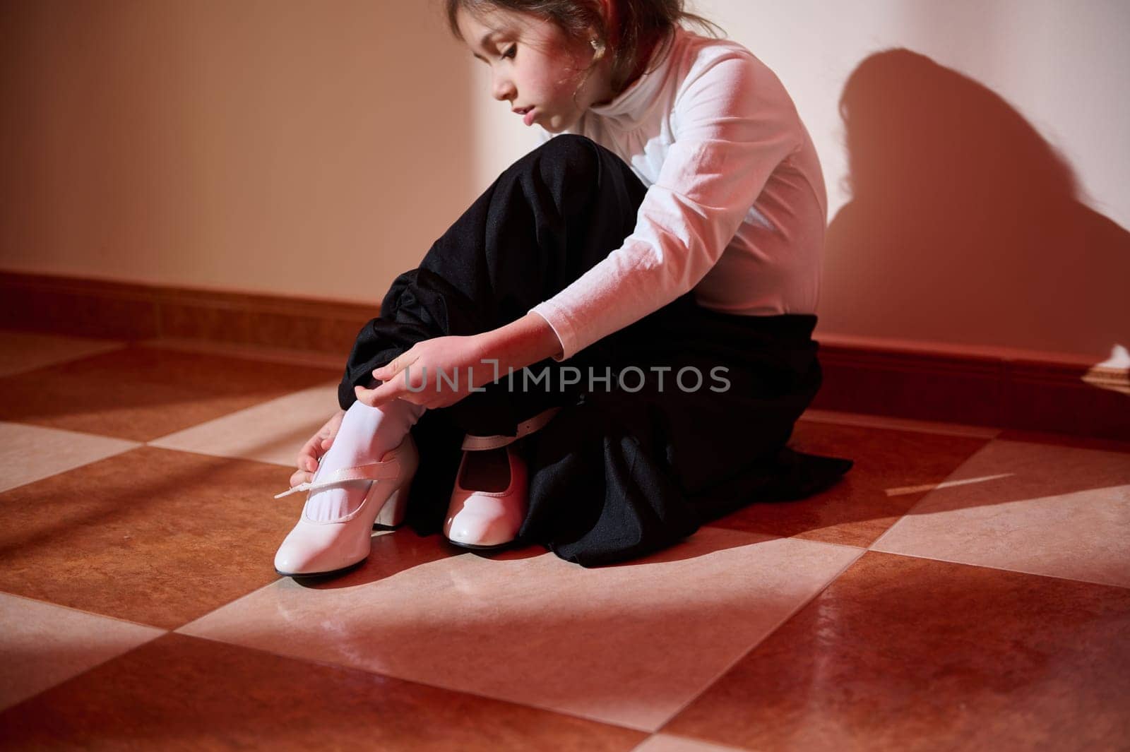 A young girl in elegant attire focuses on fastening her white shoes. The warm sunlight creates a relaxing atmosphere.