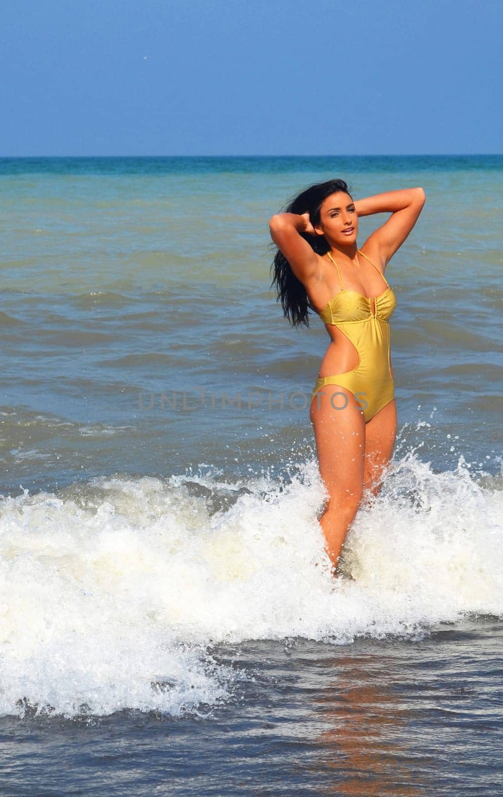 Beautiful young woman standing in a beige swimsuit in the water on the.beach in a white wave of water with her long black hair.