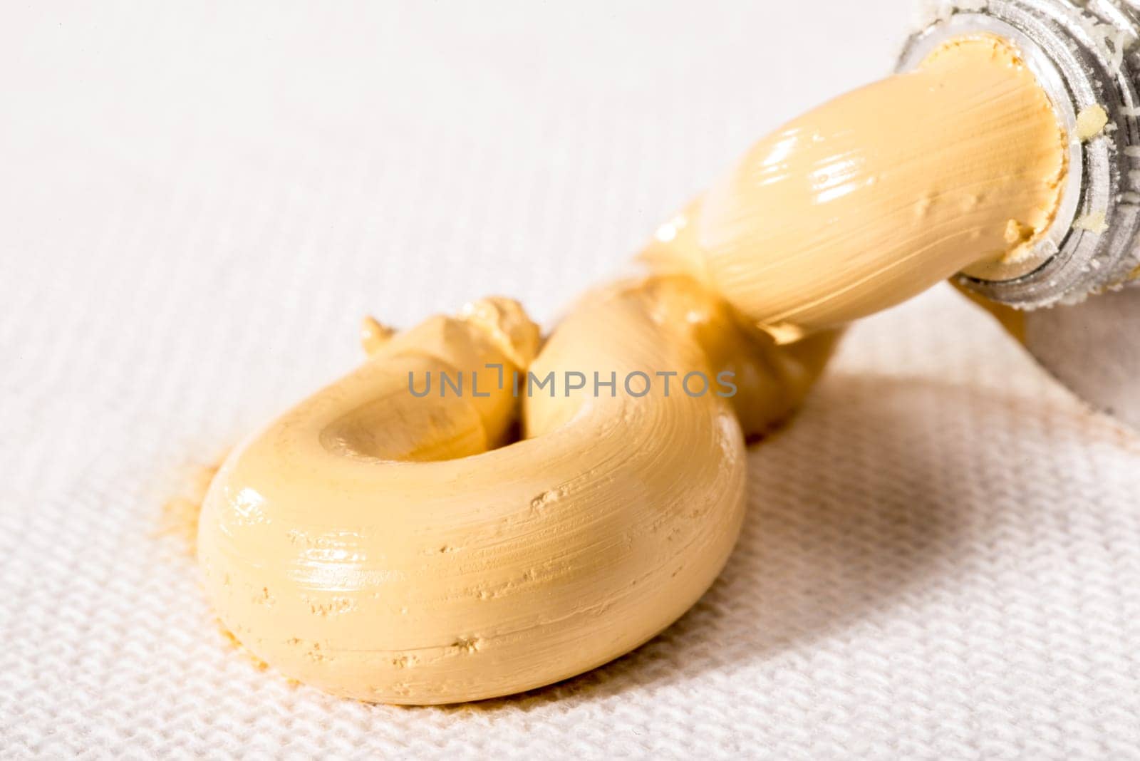 A paint tube of Naples Yellow Hue oil paint is pressed and the paste of the paint is squeezed out on to a white artist's canvas.
