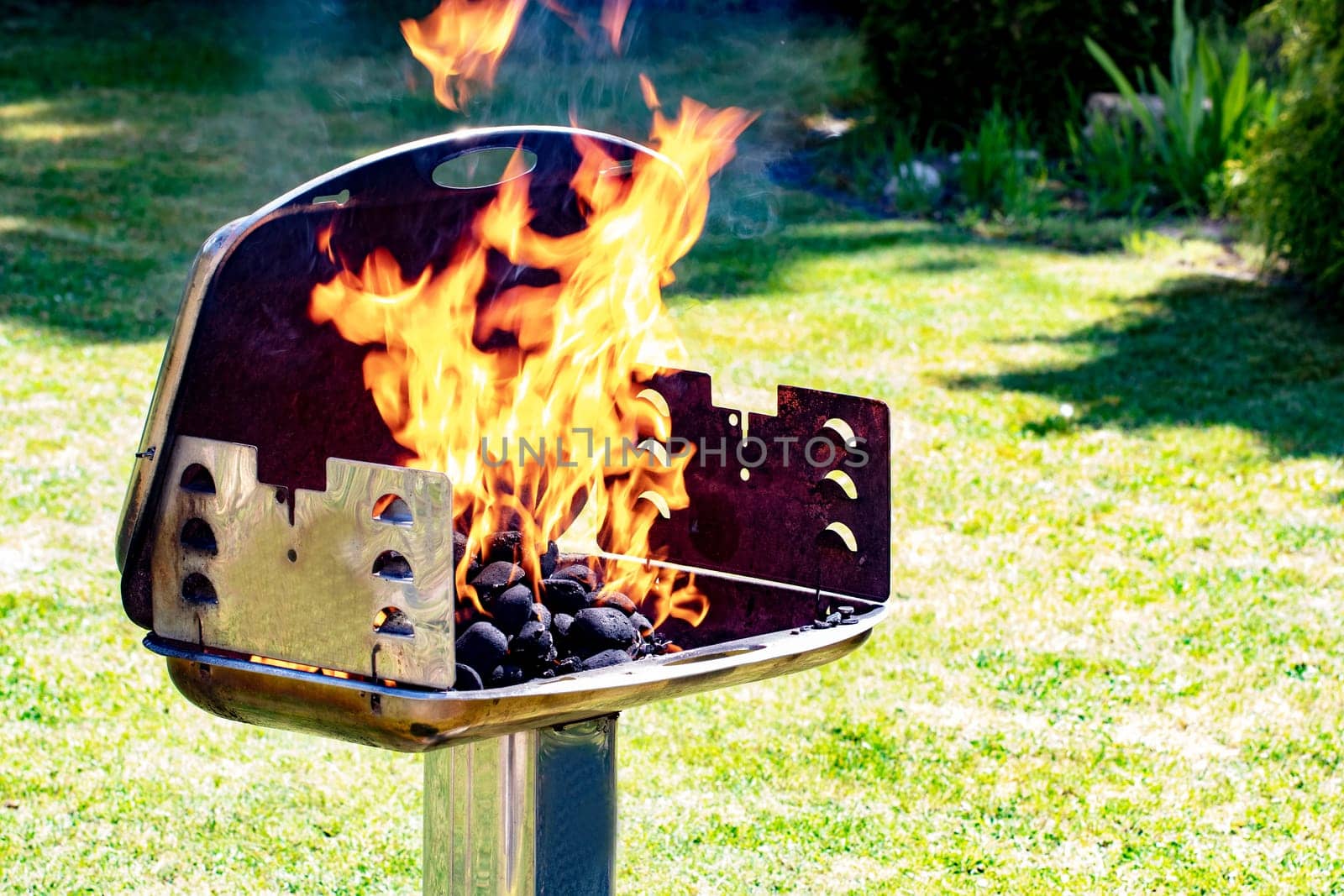 Close-up of the frozen moment of the blazing flames in a charcoal grill with green grass background of a garden. Prepearing of a barbecue party at a sunny spring day. BBQ concept. Macro.