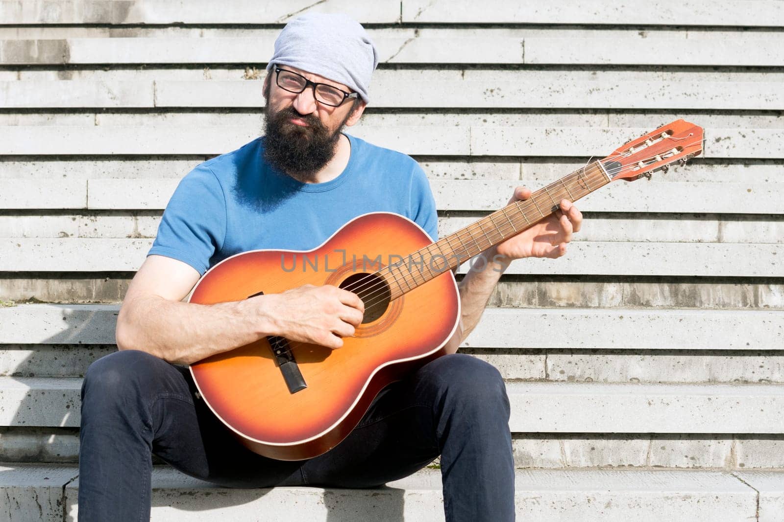 The funny bearded man with grimace is playing guitar and sitting on stairs.