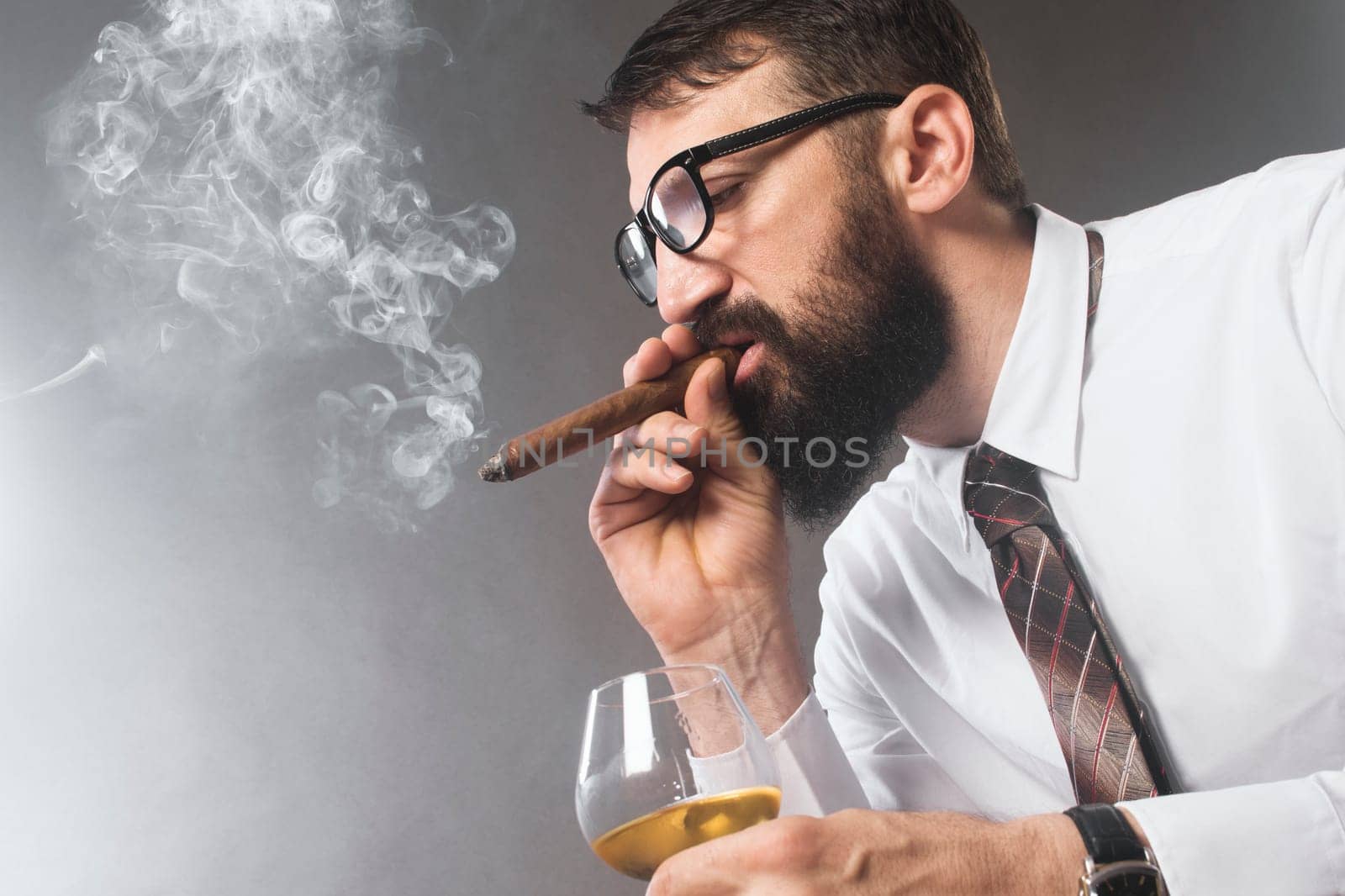 The elegant bearded man is smoking cigar and drinking alcohol on gray background.