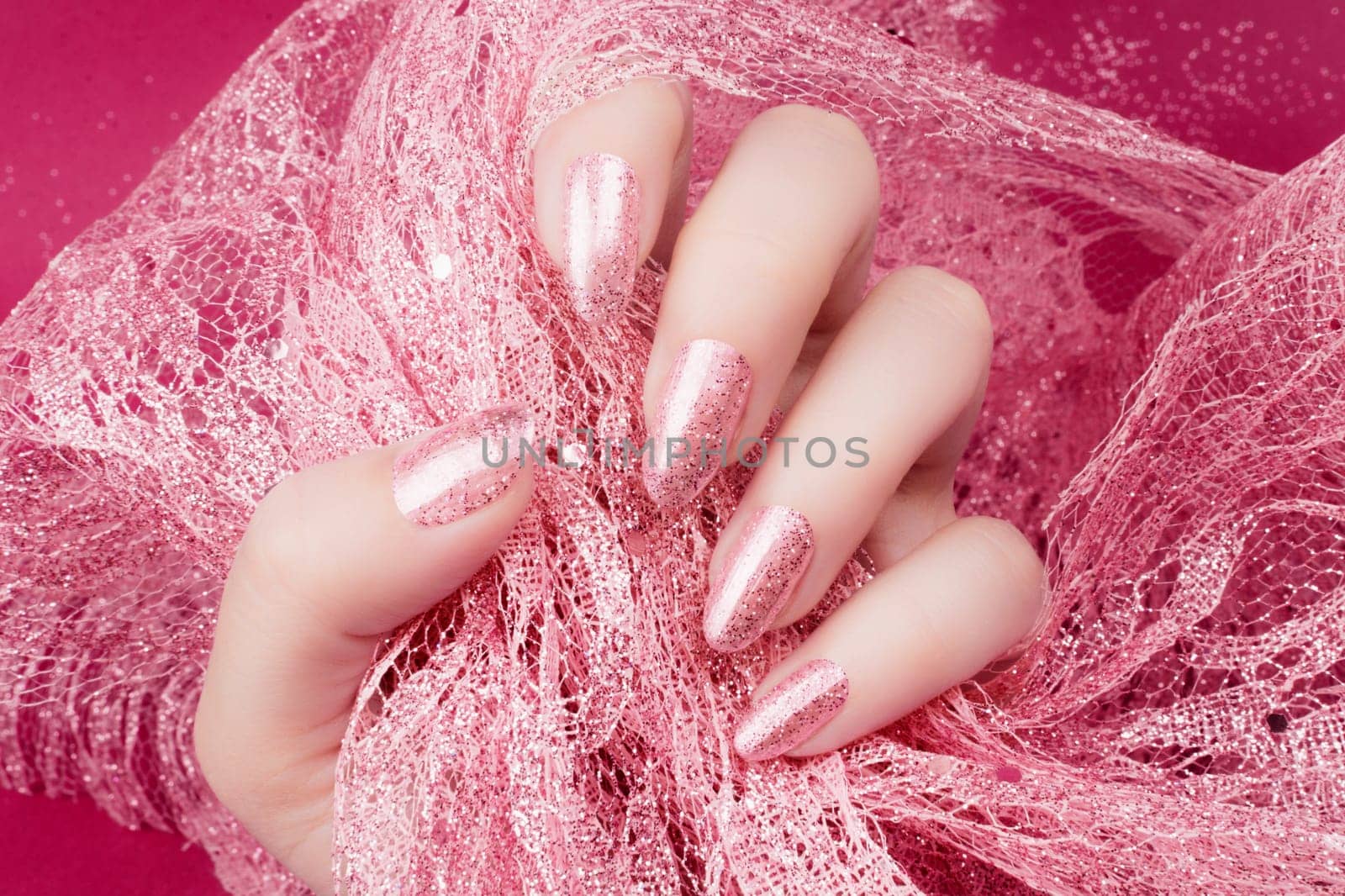 Female hand with glittered rose nails manicure is holding rose decoration on pink background.