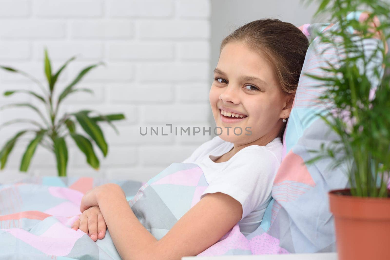 Happy ten-year-old girl in the morning lies in bed and smiles into the frame.