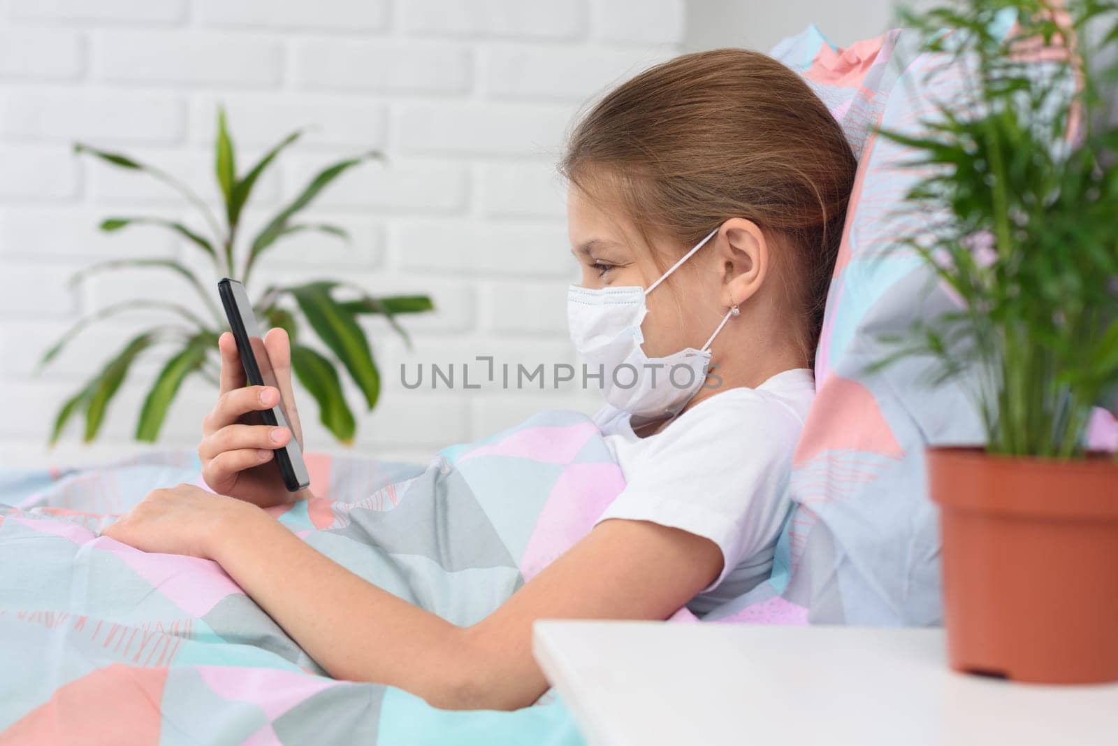 A girl in a hospital ward lies on a bed in a medical mask and does web surfing on the phone