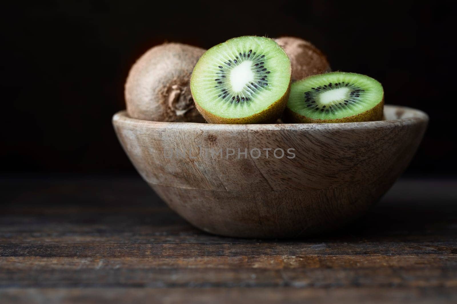 Fresh kiwis in wooden bowl on dark wooden table with dark background.