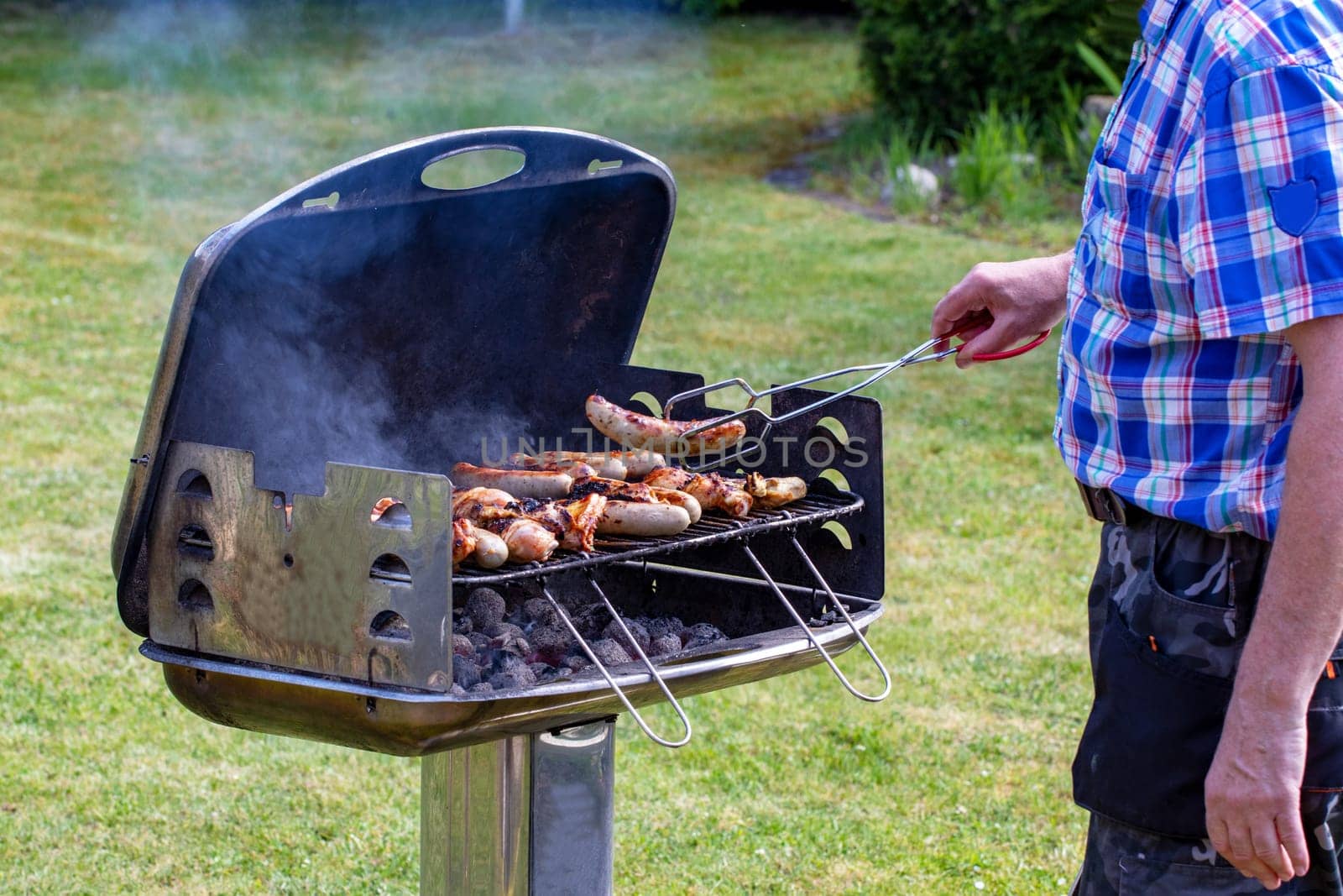 A man at the charcoal grill is grilling steaks, grill sausages and chicken wings and using barbecue tongs. Selective focus on the grill against a green lawn background on a sunny spring day. BBQ concept. Macro.