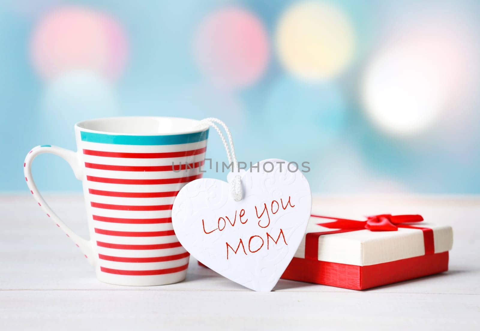 Mothers day greeting card. Decorative composition of a mug with gift or present box and a tag in shape of a heart with inscription Love you mom on a bright table over blurred background.