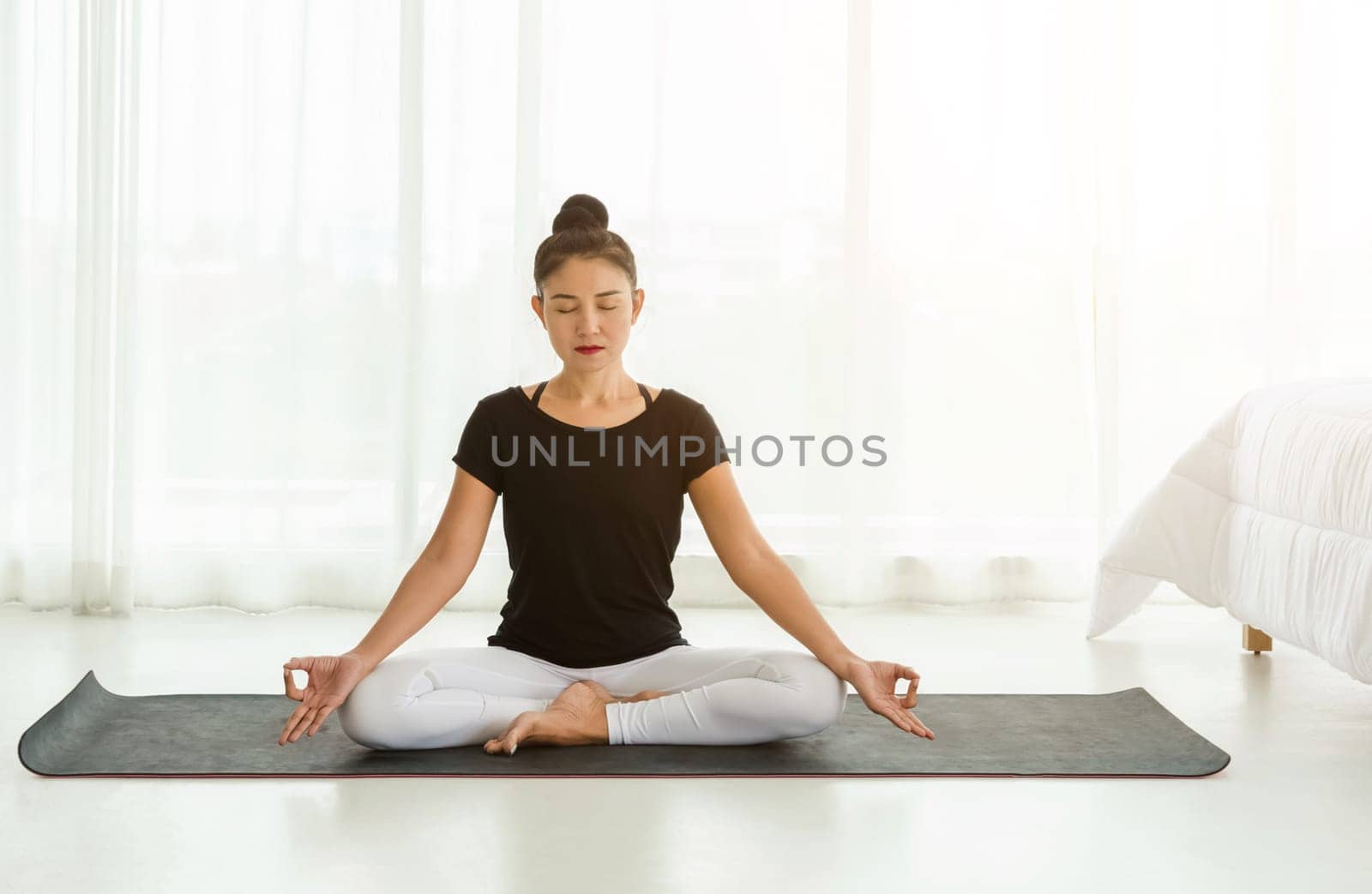 Middle aged women practicing yoga in Easy seat pose or Sukhasana with with mudra. Meditation with yoga in white bedroom after wake up in the morning. Concept of exercise, relaxation and healthcare.
