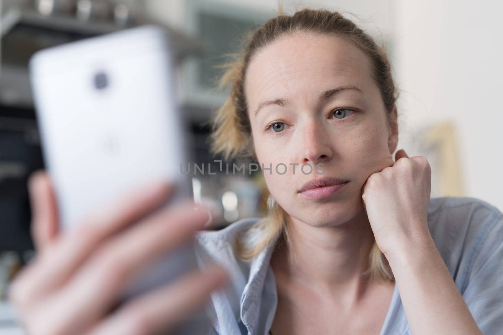 Worried woman indoors at home kitchen using social media apps on phone for video chatting and stying connected with her loved ones. Stay at home, social distancing lifestyle.