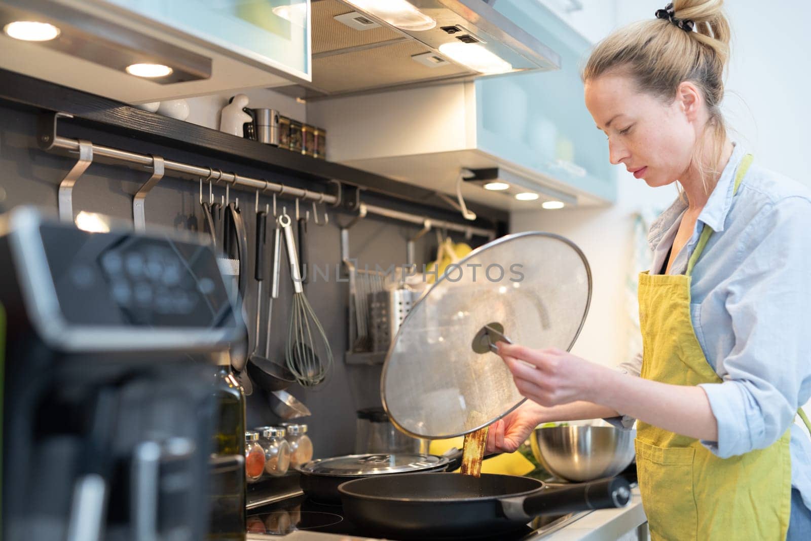 Stay at home housewife woman cooking in kitchen, stir frying dish in a saucepan, preparing food for family dinner. Blonde caucasian model in her 30is, real cooking not looking in camera.