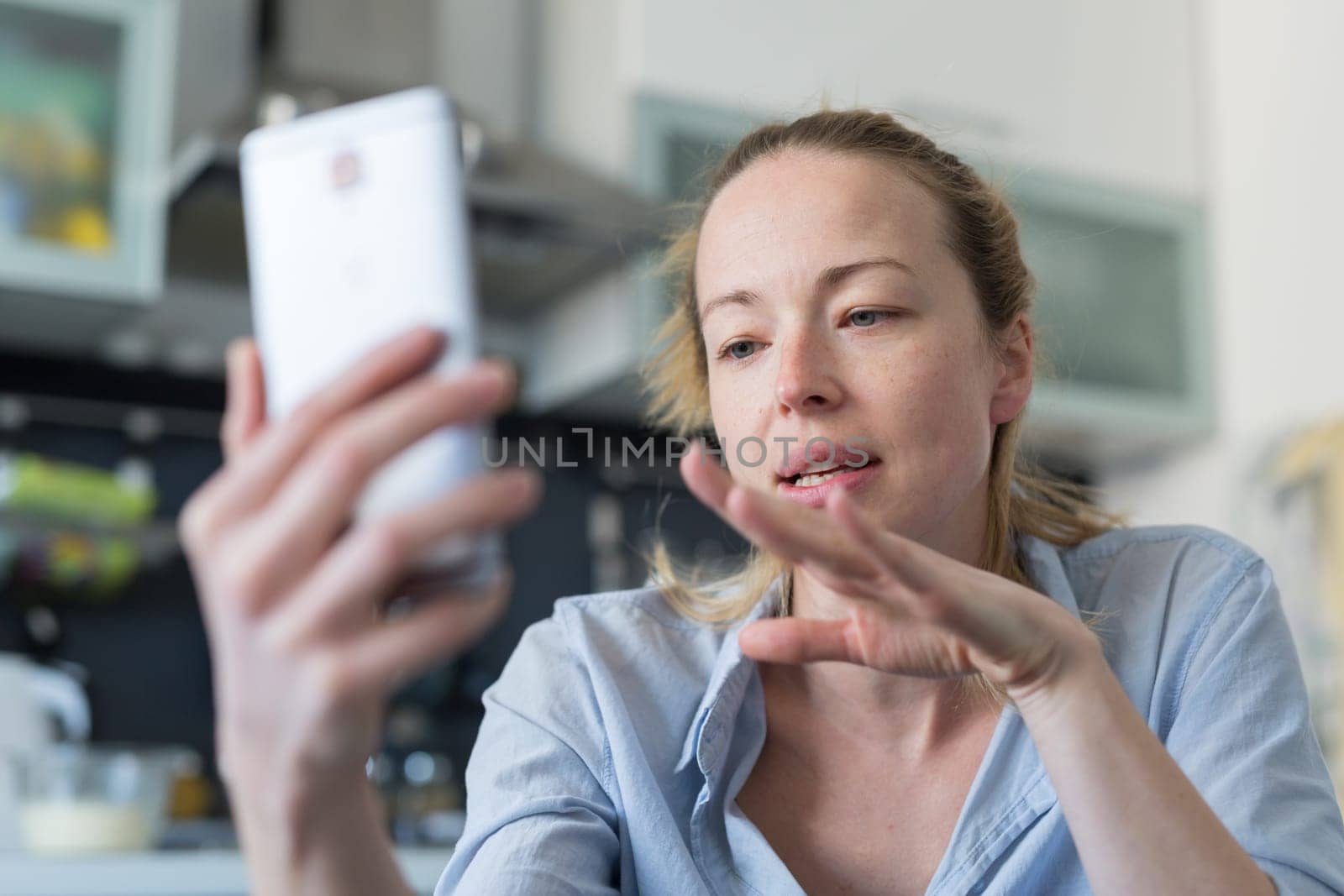 Young woman indoors at home kitchen using social media apps on phone for video chatting and stying connected with her loved ones. Stay at home, social distancing lifestyle.