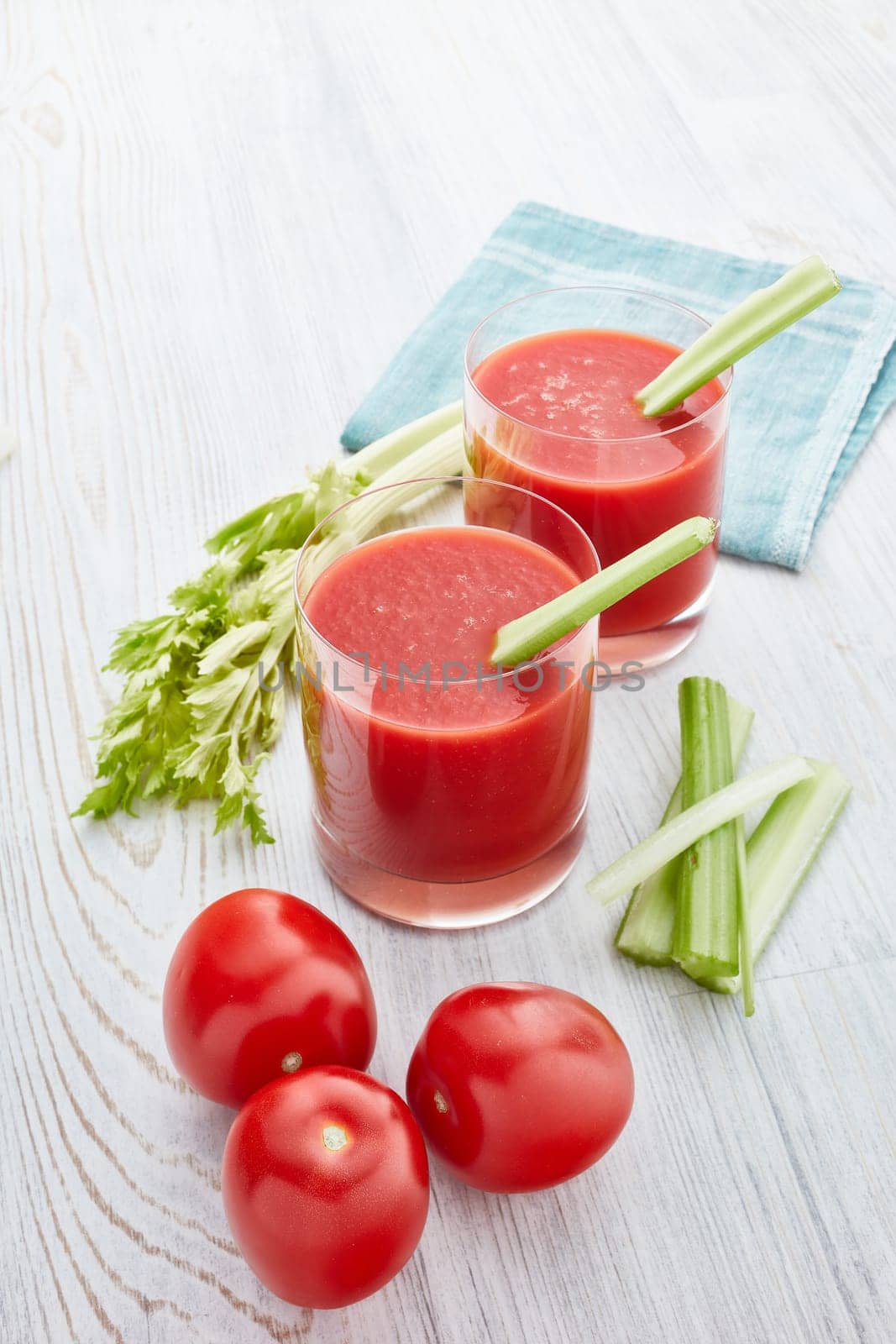 Two glasses with fresh tomato juice, celery, parsley and ripe tomatoes on light wooden background. Top view