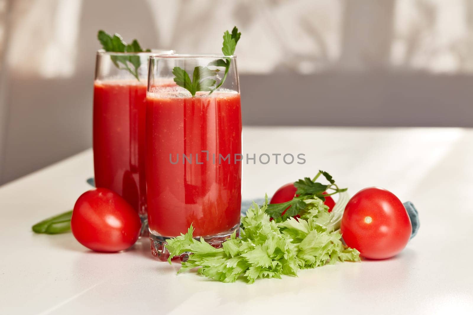Two glasses with fresh tomato juice, celery and ripe tomatoes on white table near window. With copy space