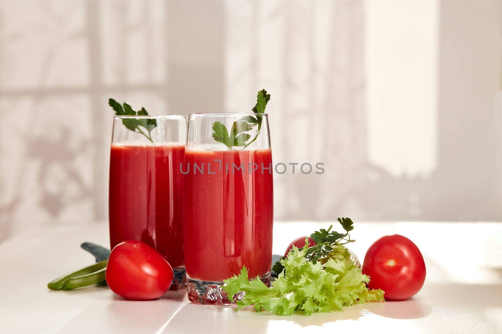 Two glasses with fresh tomato juice, celery and ripe tomatoes on white table near window. With copy space