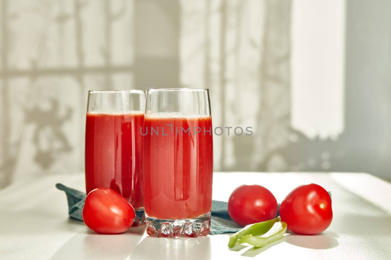 Two glasses with fresh tomato juice, celery and ripe tomatoes on white table near window. With copy space