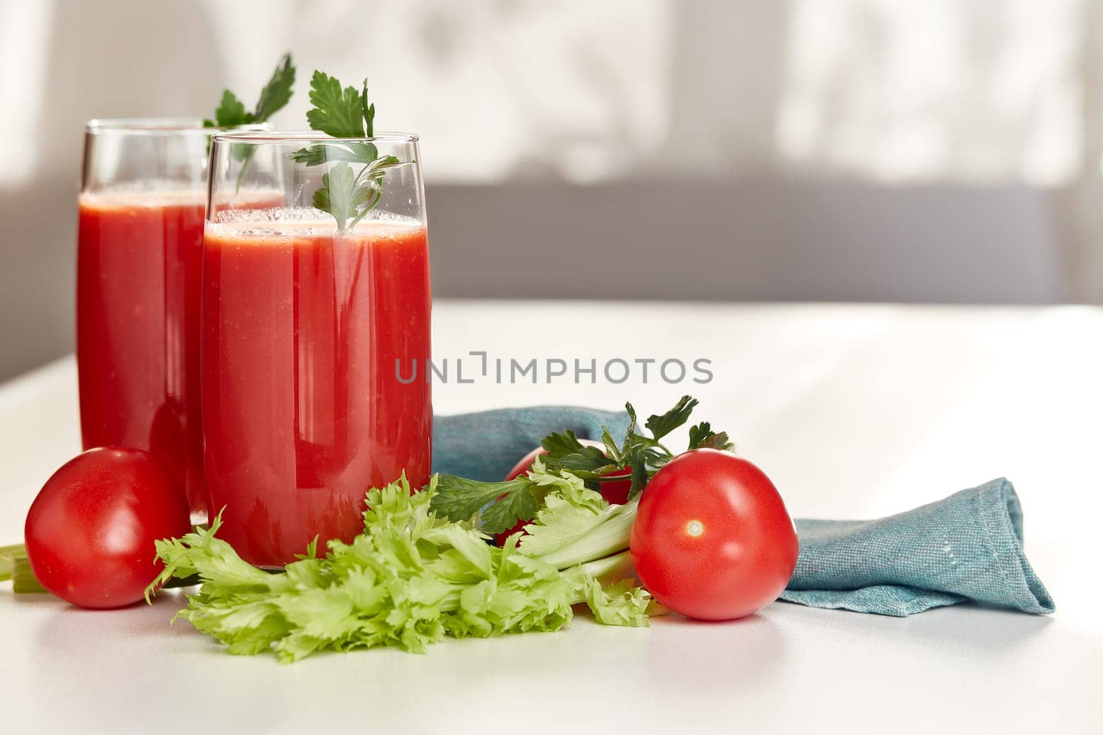 Two glasses with fresh tomato juice, celery and ripe tomatoes on white table near window. With copy space