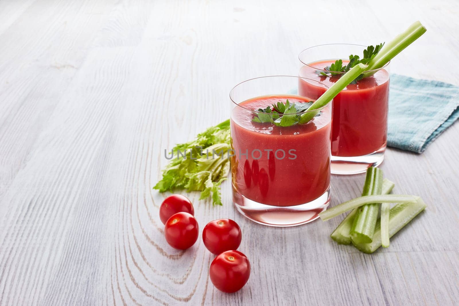 Two glasses with fresh tomato juice, celery, parsley and ripe tomatoes on light wooden background. Top view