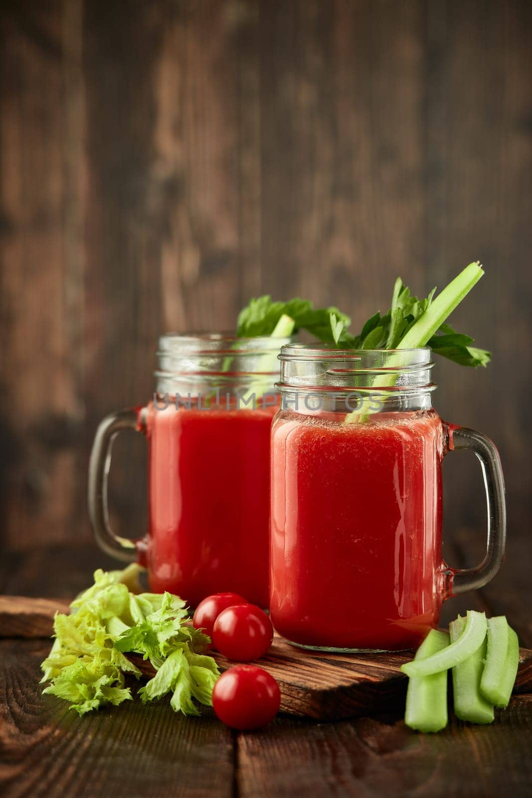 Two glasses with fresh tomato juice, celery, parsley and ripe tomatoes on dark brown wooden background. Healthy beverage, vegetable juice