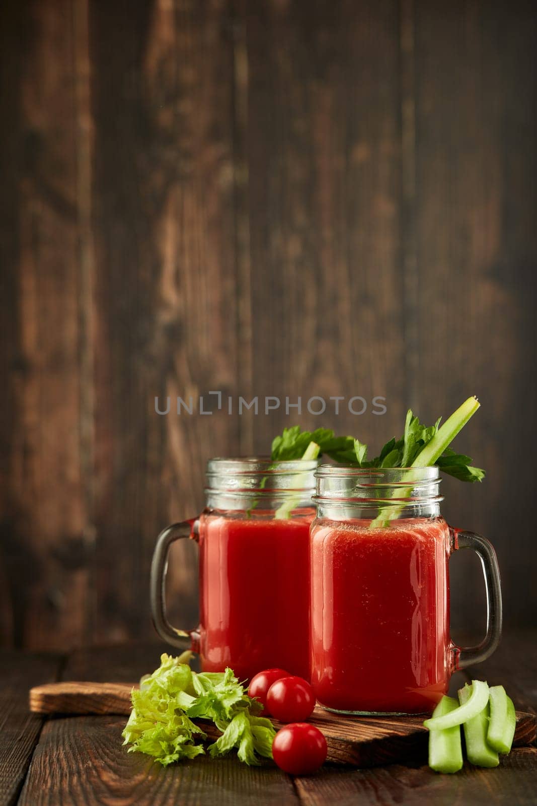 Two glasses with fresh tomato juice, celery, parsley and ripe tomatoes on dark brown wooden background. Healthy beverage, vegetable juice
