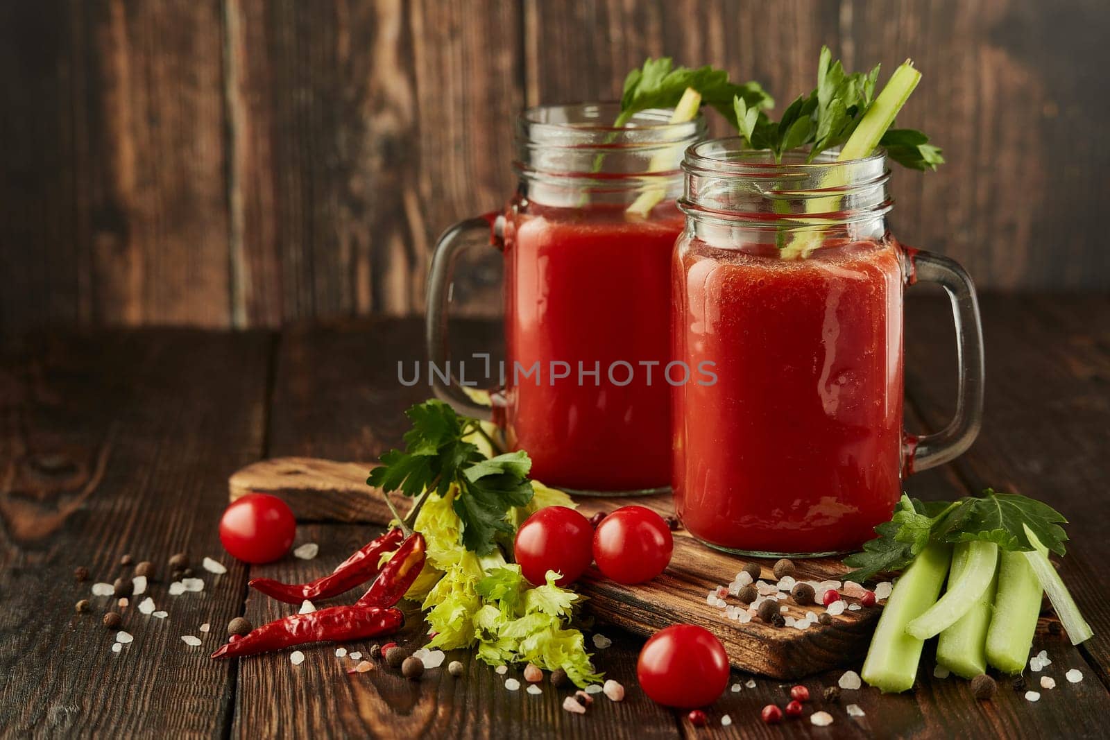 Two glasses with fresh tomato juice, celery, parsley, spices and ripe tomatoes on dark brown wooden background with copy space. Healthy beverage, vegetable juice