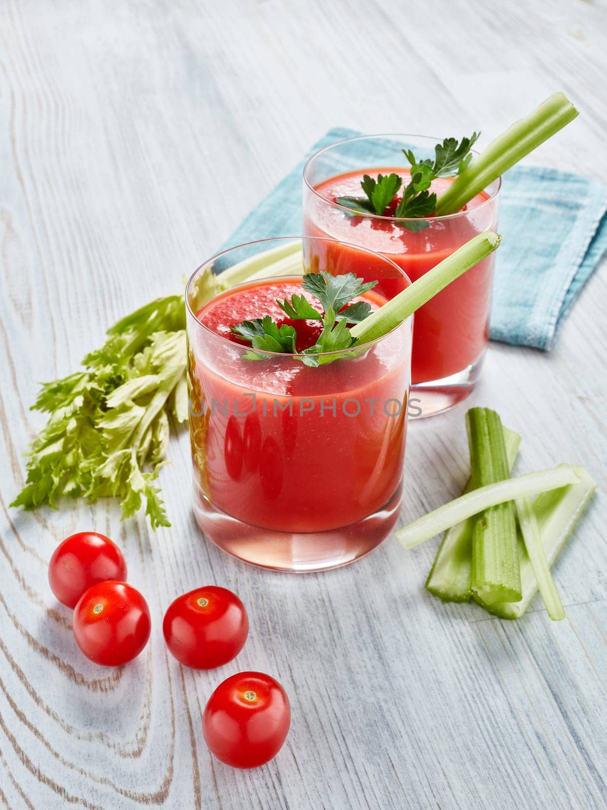 Two glasses with fresh tomato juice, celery, parsley and ripe tomatoes on light wooden background. Top view