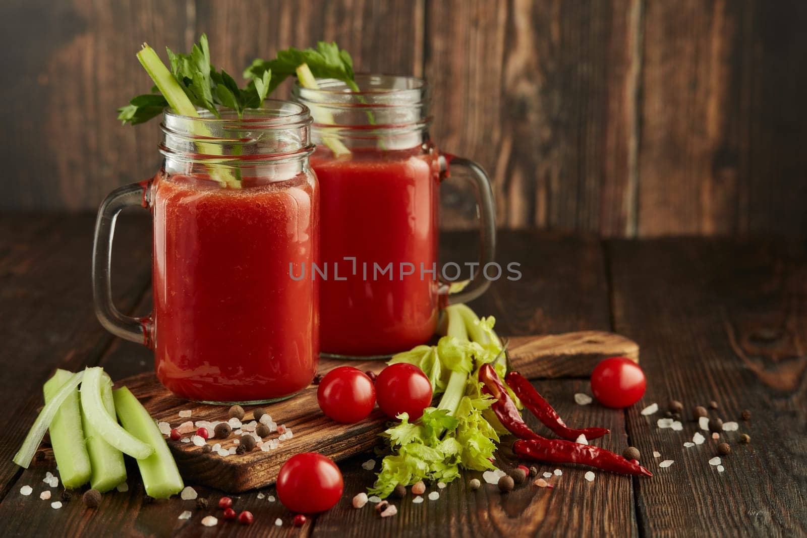 Two glasses with fresh tomato juice, celery, parsley, spices and ripe tomatoes on dark brown wooden background with copy space. Healthy beverage, vegetable juice