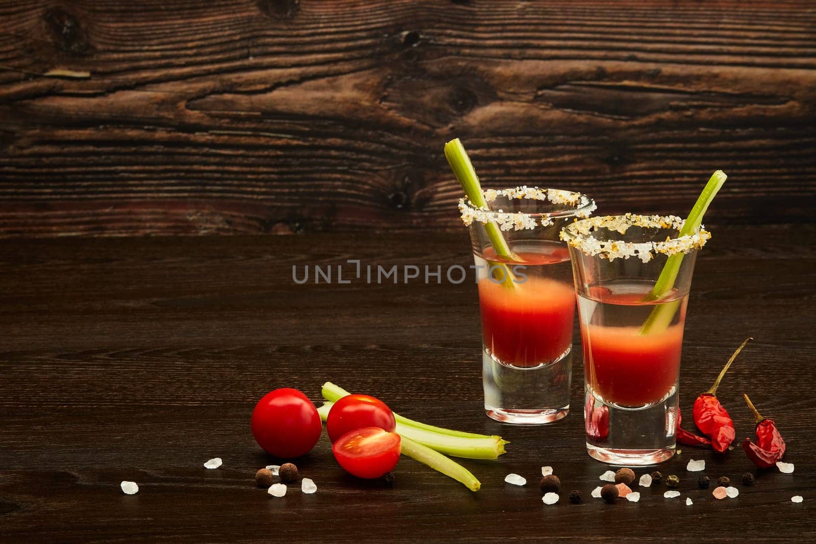 Two shot glasses with cocktail Bloody Mary, celery, spices and ripe tomatoes on dark wooden background with copy space