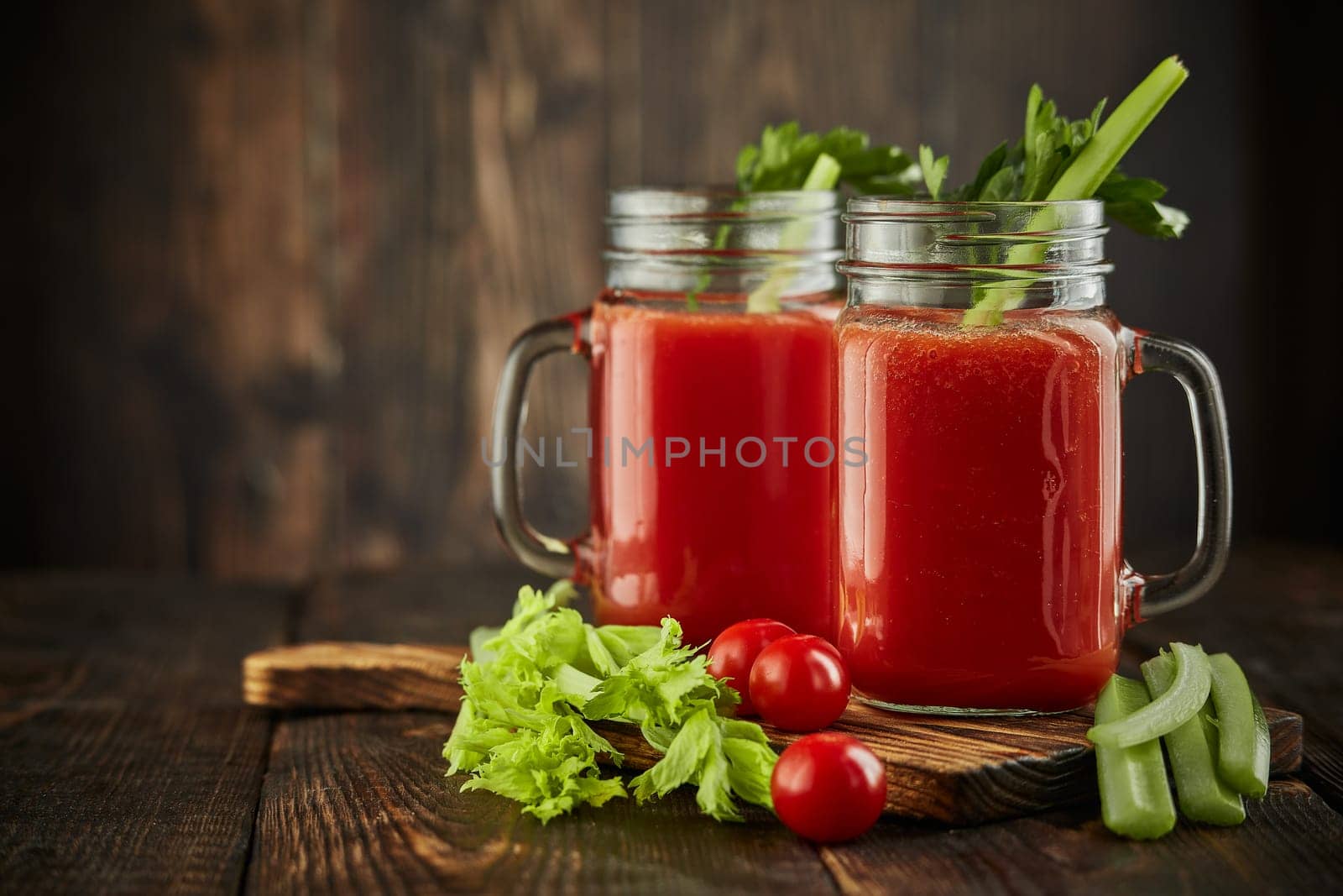 Two glasses with fresh tomato juice, celery, parsley and ripe tomatoes on dark brown wooden background with copy space. Healthy beverage, vegetable juice