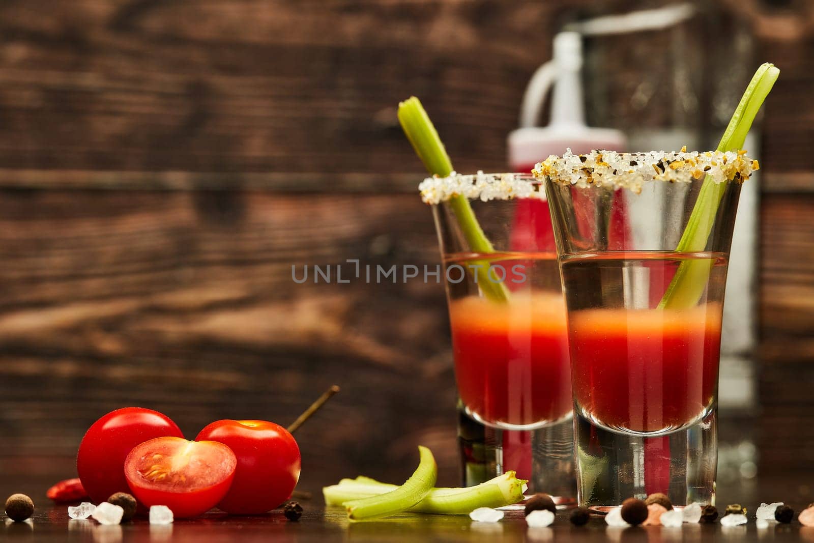 Two shot glasses with cocktail Bloody Mary, celery, spices and ripe tomatoes on dark wooden background with copy space
