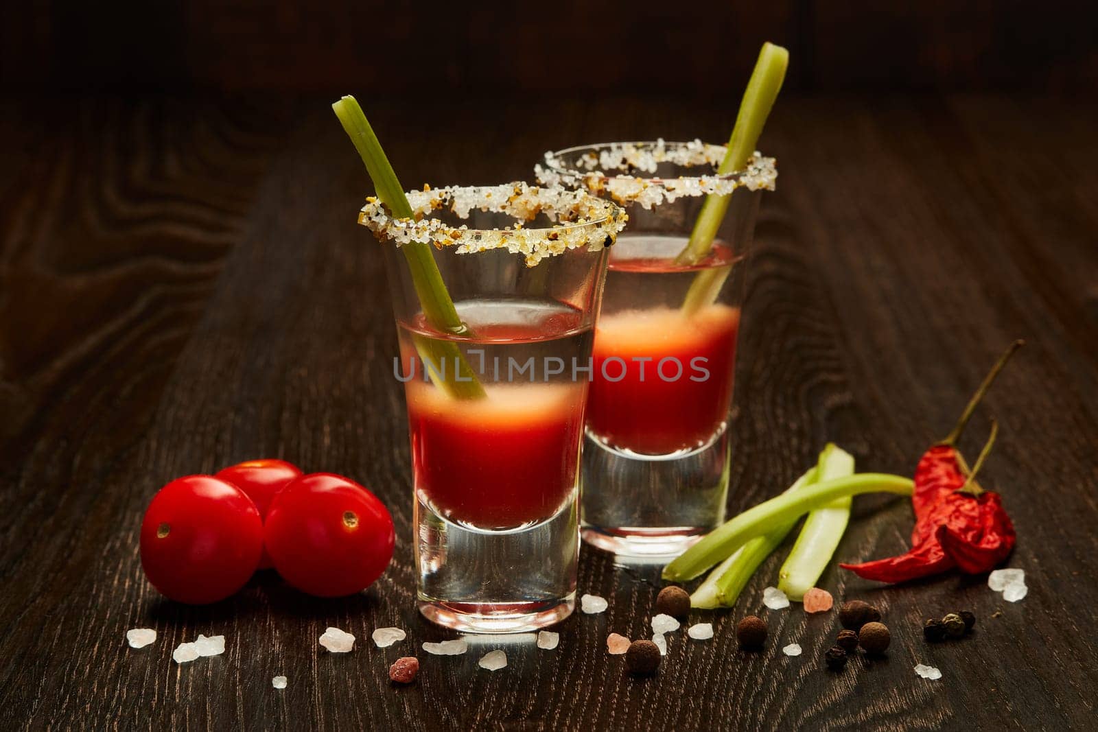 Two shot glasses with cocktail Bloody Mary, celery, spices and ripe tomatoes on dark wooden background