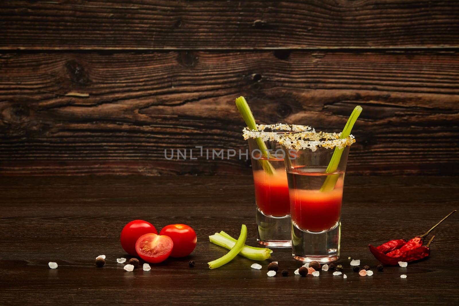 Two shot glasses with cocktail Bloody Mary, celery, spices and ripe tomatoes on dark wooden background with copy space