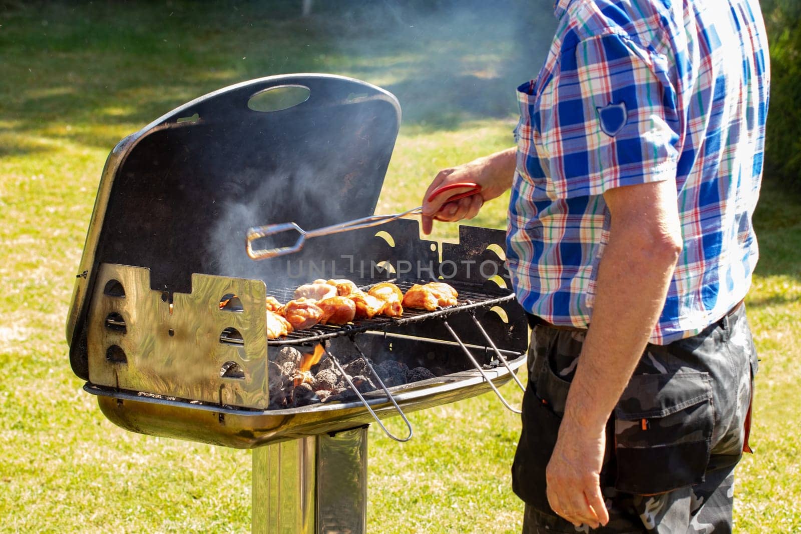 A man at the charcoal grill uses grill tongs to place the already seasoned raw meat on the grill. Selective focus on the grill against a green lawn background on a sunny spring day. BBQ concept. Macro.