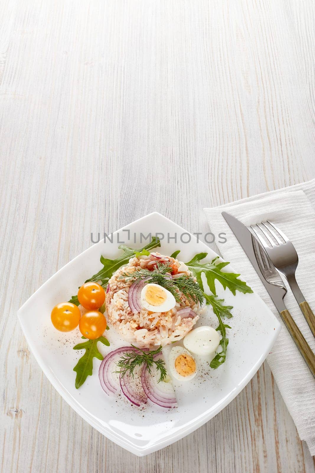 Tuna salad with  rice, arugula and tomatoes on white plate on light wooden background. Top view with copy space. Healthy food, seafood menu, Setting table