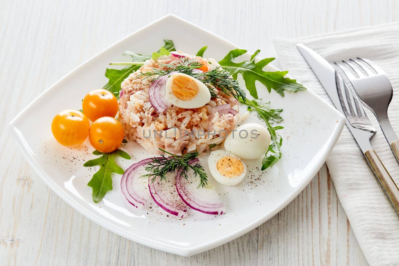 Tuna salad with  rice, arugula and tomatoes on white plate on light wooden background. Top view. Healthy food, seafood menu, Setting table