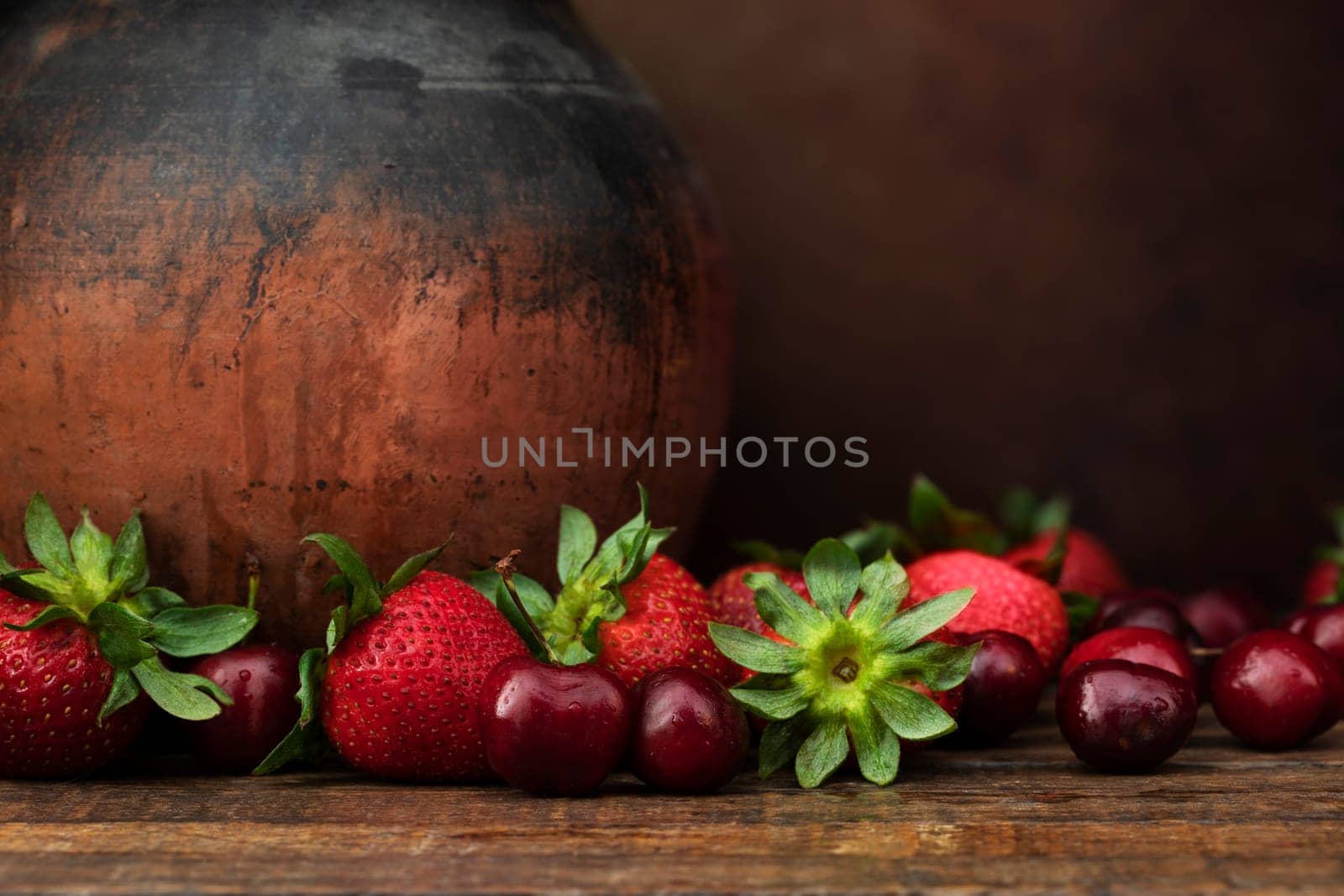 Strawberries and cherries on dark wooden table with rustic pottery jug and copy space.