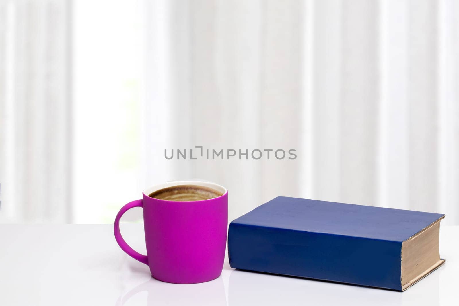Books backgrounds. Closeup of pink coffee mug and blue book on white desk in front of abstract blurred curtain background. Morning coffee on a sunny day.