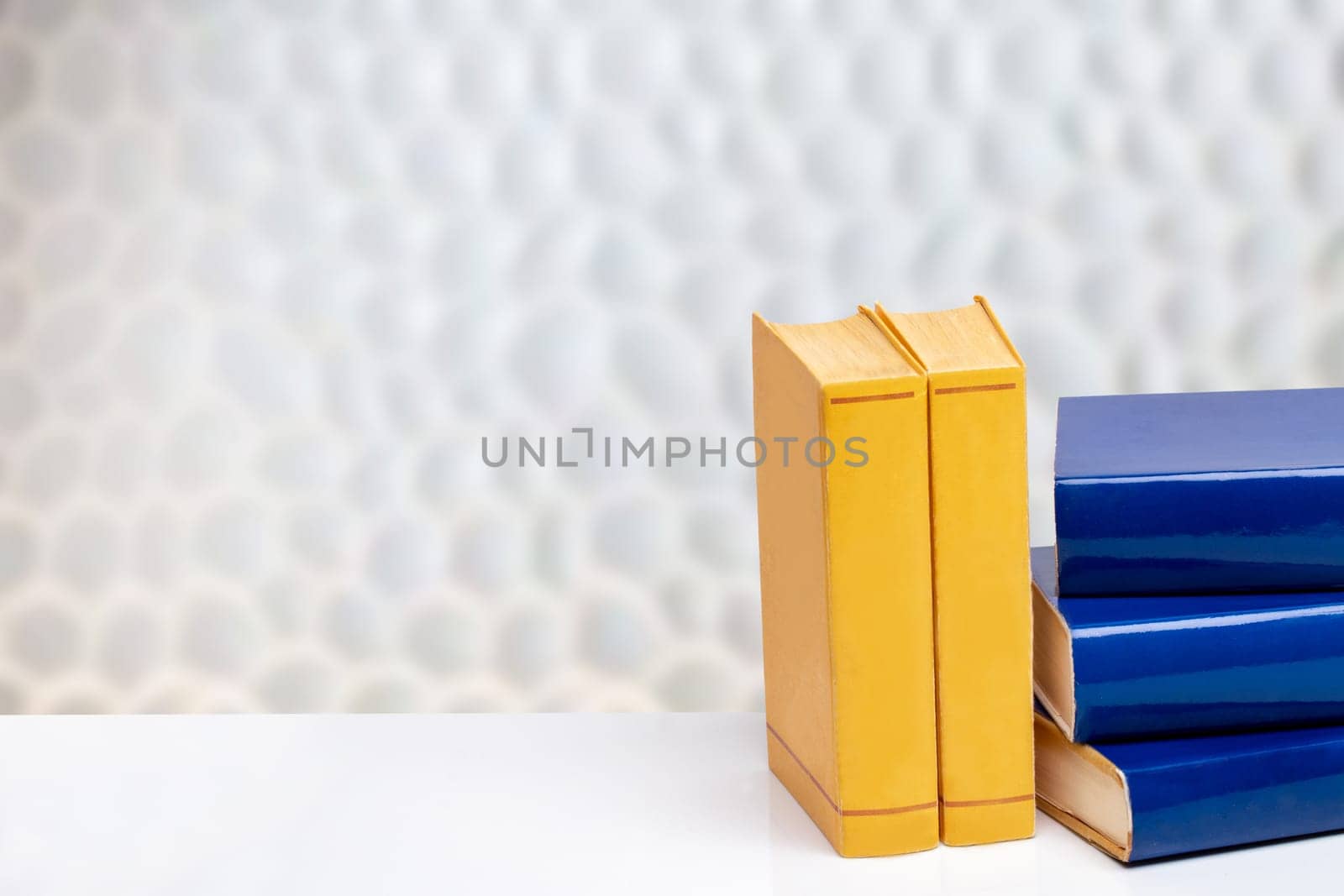 Books backgrounds. Close-up of a stack blue and yellow books on a white table in front of abstract blurred bright background. Space for text and montage.