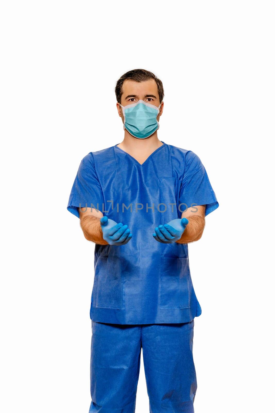 Male doctor in scrubs with protective face mask and gloves, reaching out with open hands on plain white background. Coronavirus or covid 19 concept