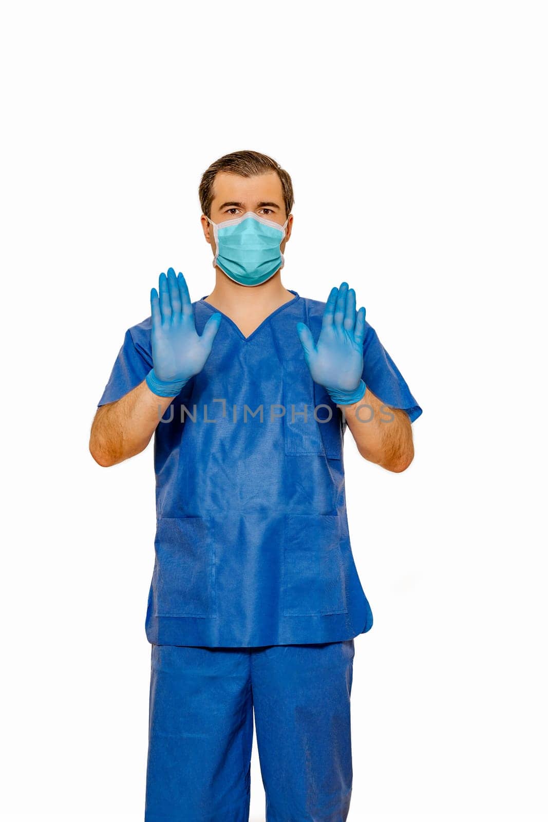 Male doctor in scrubs with protective face mask and gloves showing stop sign with both his hands and palms. Plain white background Coronavirus concept