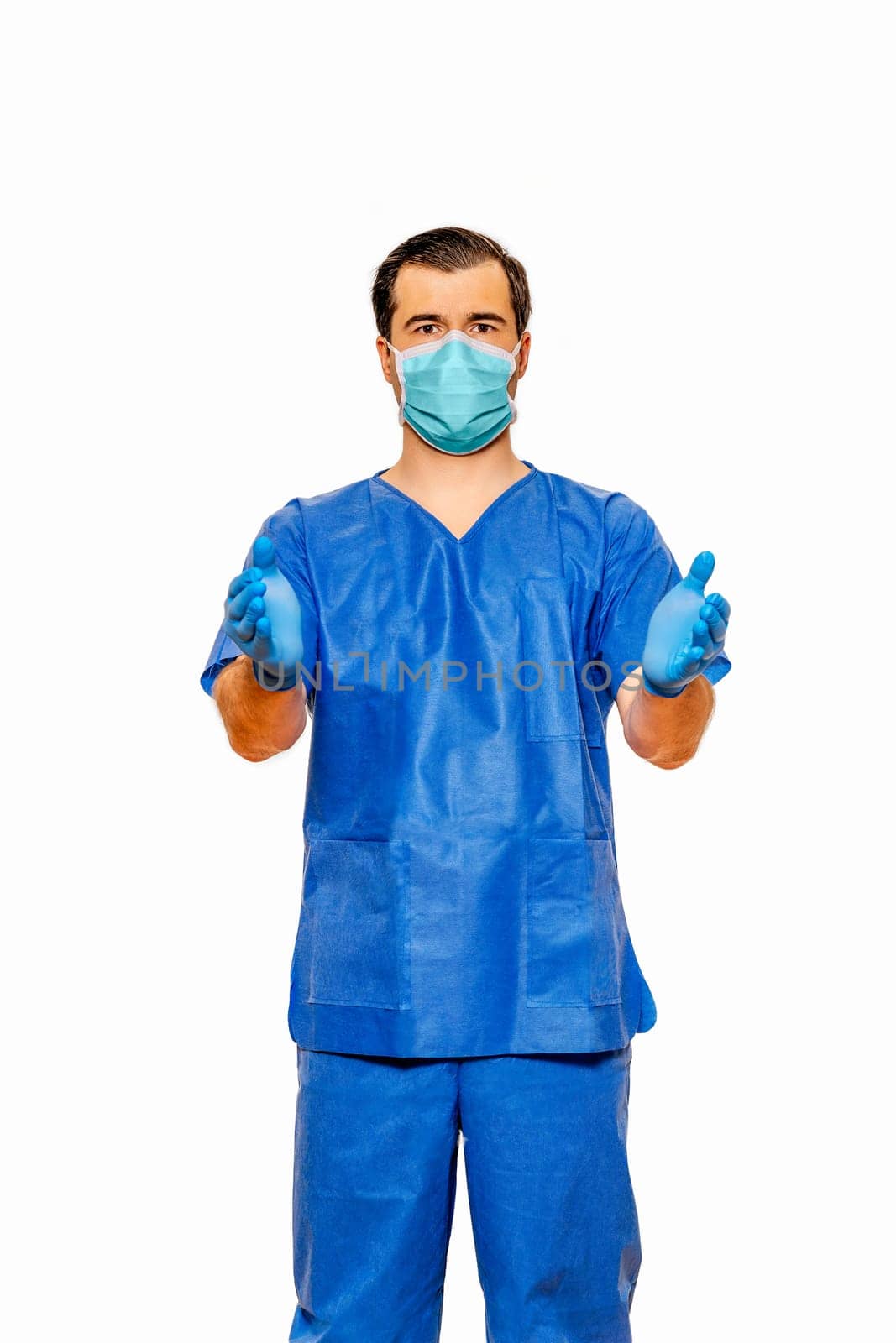 Male doctor in scrubs with protective face mask and gloves, inviting with open hands on plain white background. Coronavirus or covid 19 concept