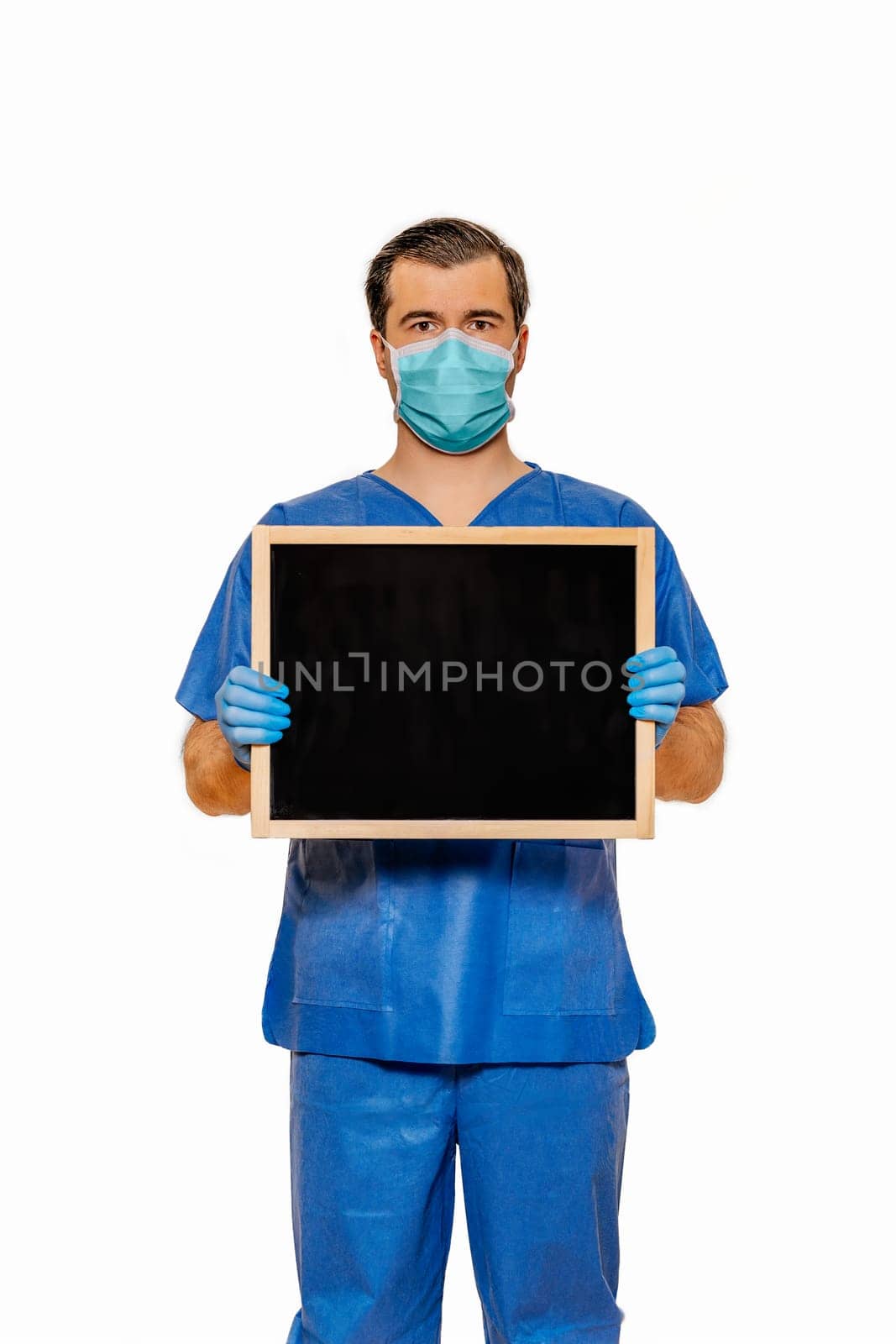 Male doctor in scrubs with protective face mask holding clean blackboard with space for your text and ideas Plain white background Coronavirus concept