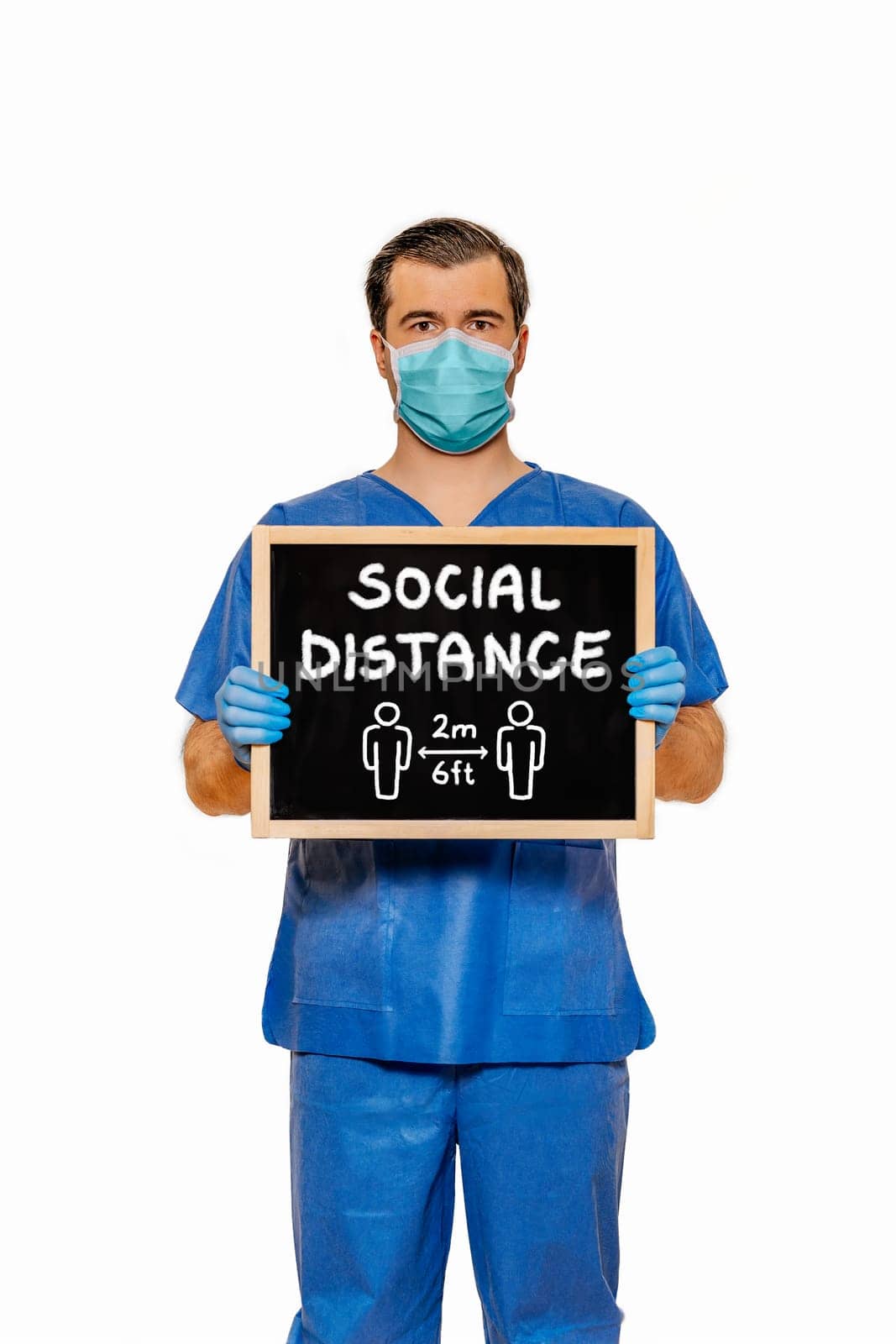 Male doctor in scrubs with protective face mask, holding blackboard with chalk text Social Distance. Plain white background. Coronavirus concept