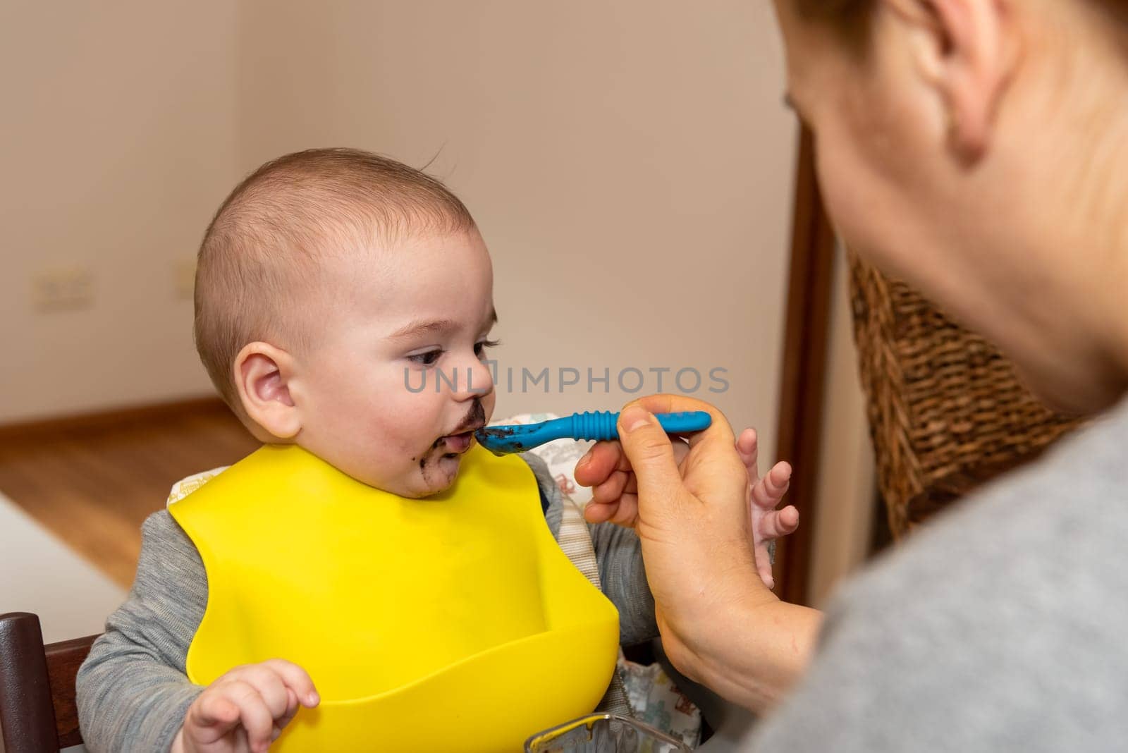 Adorable little baby boy in feeding chair being spoon fed by his mother