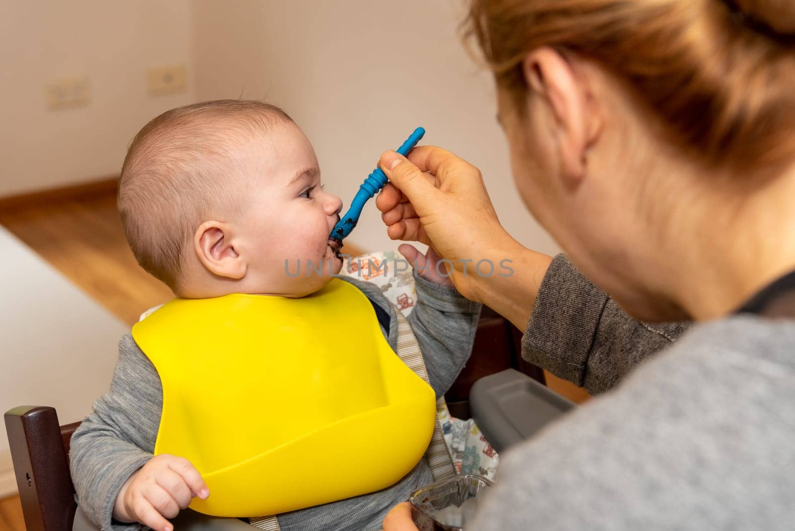 Adorable little baby boy in feeding chair being spoon fed by his mother