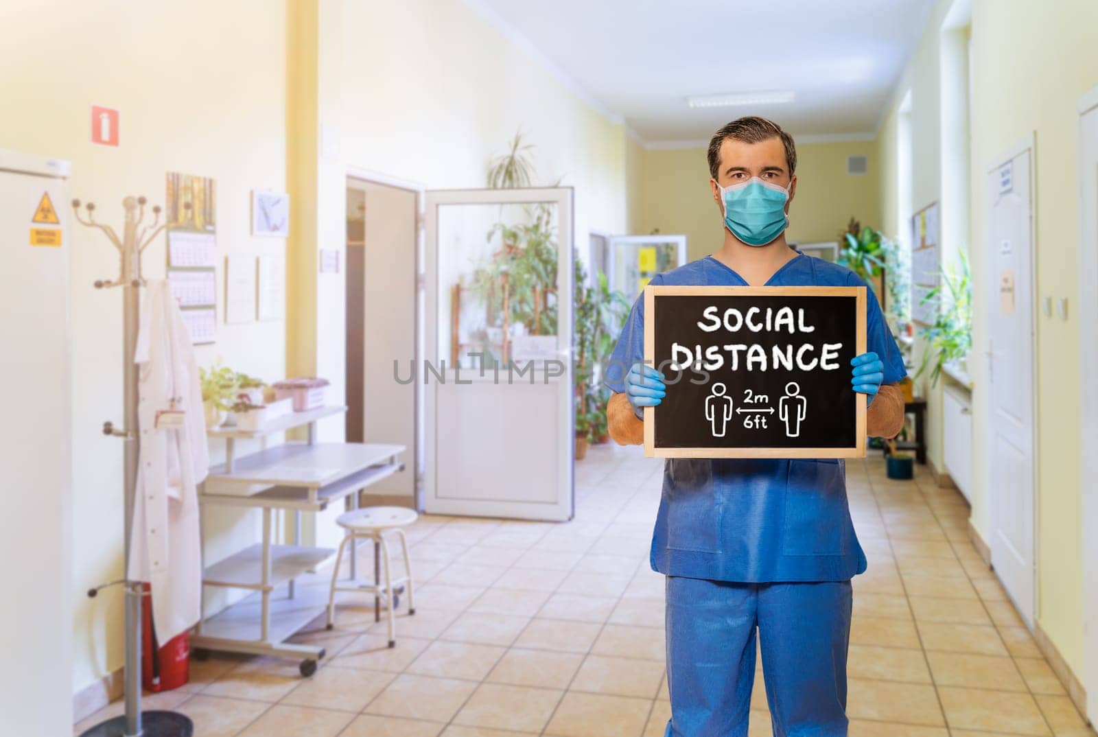 Male doctor in scrubs with protective face mask, holding blackboard with chalk handwriting Social Distance on defocused hospital corridor background