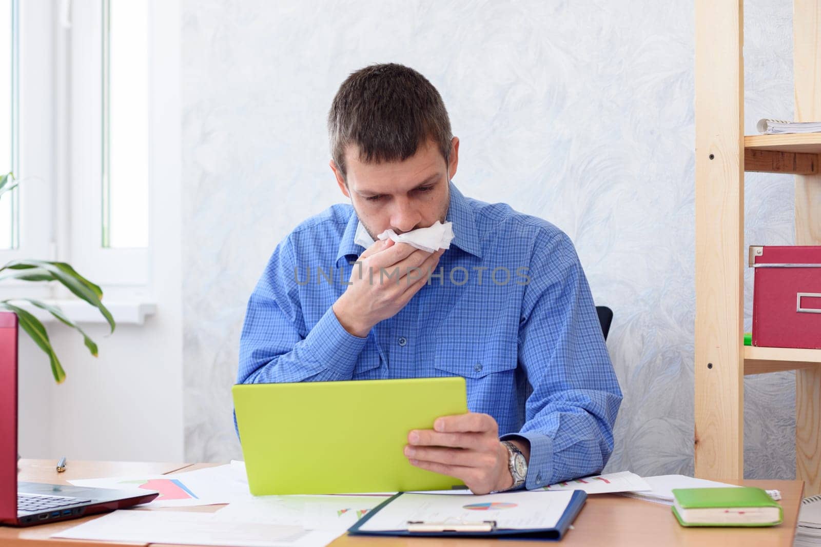 A man in the office with a tablet in his hand coughs in a napkin