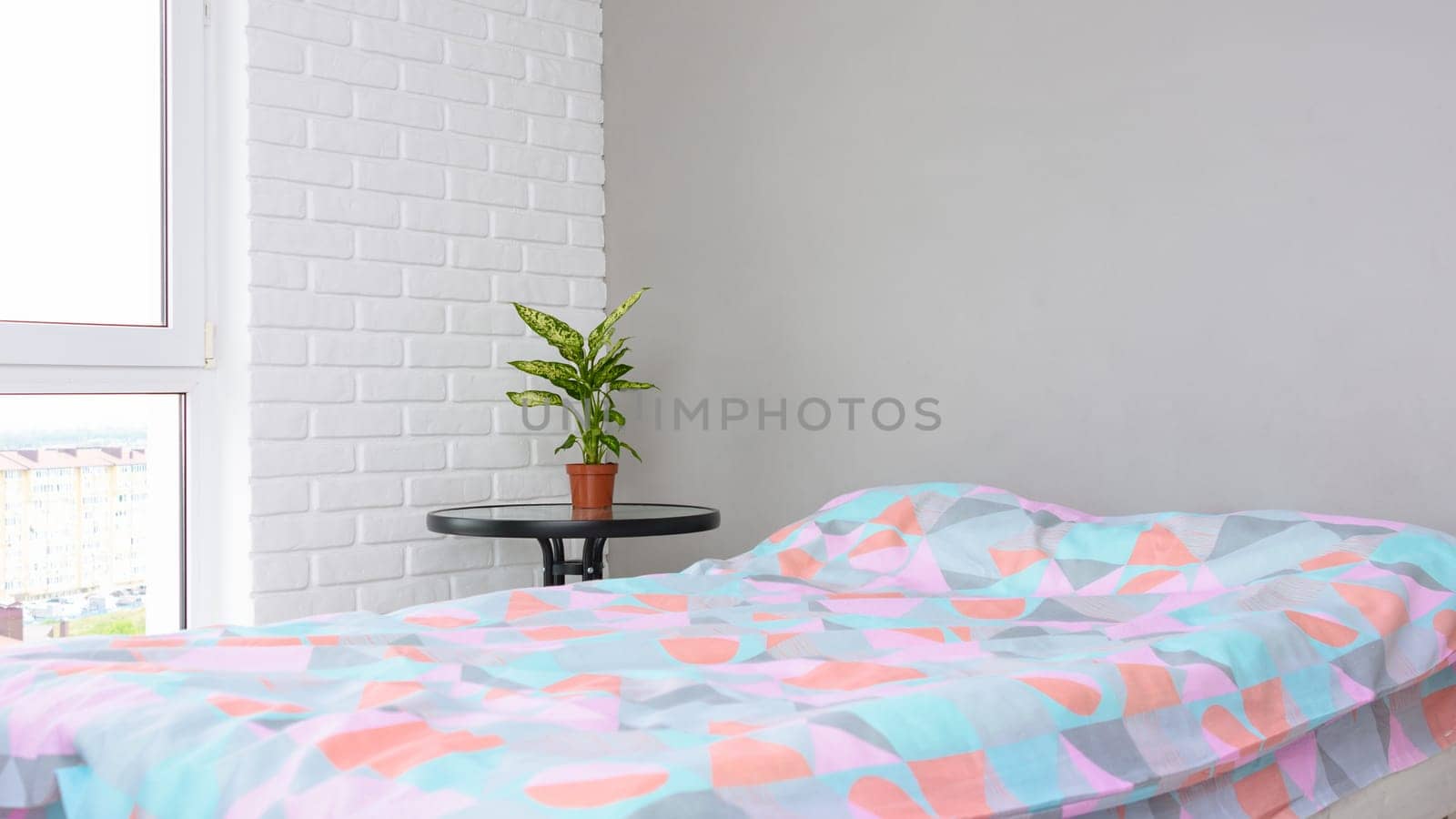 A fragment of the bedroom interior, a large made-up bed by the window, next to it is a bedside table with a flower